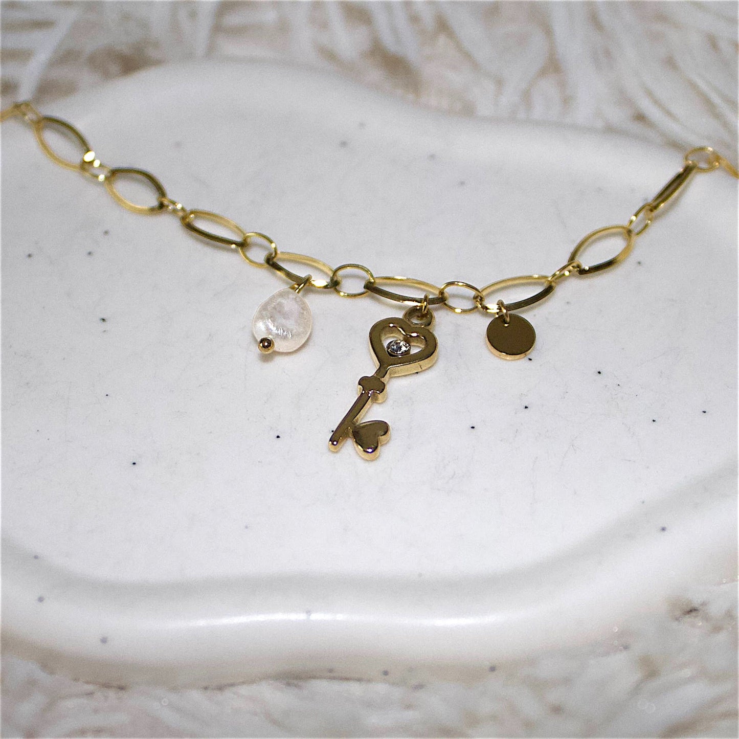 Stylish waterproof bracelet made of gold-plated stainless steel, featuring a key charm and a delicate pearl detail. This durable and elegant bracelet is presented on a white stone backround, so the charms are visible.